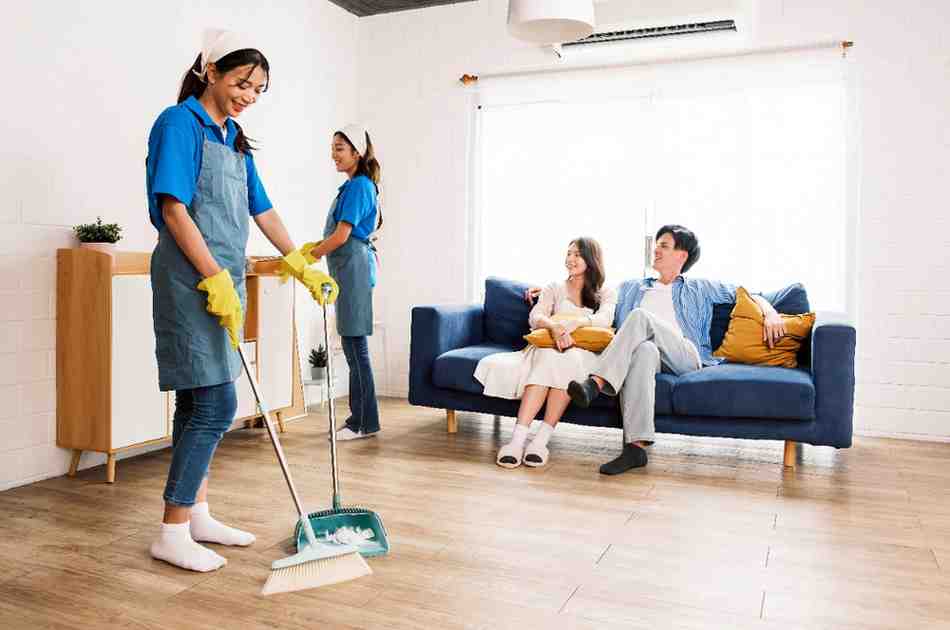 young-professional-cleaning-service-women-worker-working-house-one-sweeps-broomsticks-wooden-floor-other-cleans-low-cabinet-while-couple-owners-sit-sofa (1)